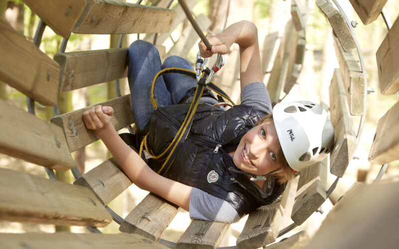 Ein Kind, das einen Helm und einen Sicherheitsgurt trägt, lächelt beim Klettern durch einen Holztunnel in einem Hochseilgarten.