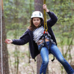 Ein Mädchen, das einen Helm und ein Gurtzeug trägt, fährt mit einer Seilrutsche durch ein bewaldetes Gebiet, mit Bäumen und Grünpflanzen im Hintergrund.