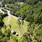 Luftaufnahme einer grasbewachsenen Lichtung inmitten eines dichten Waldes, auf der ein paar kleine Gebäude, geparkte Autos und Spielgeräte zu sehen sind.