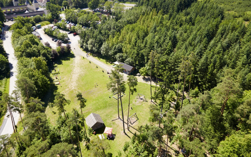 Luftaufnahme einer grasbewachsenen Lichtung inmitten eines dichten Waldes, auf der ein paar kleine Gebäude, geparkte Autos und Spielgeräte zu sehen sind.