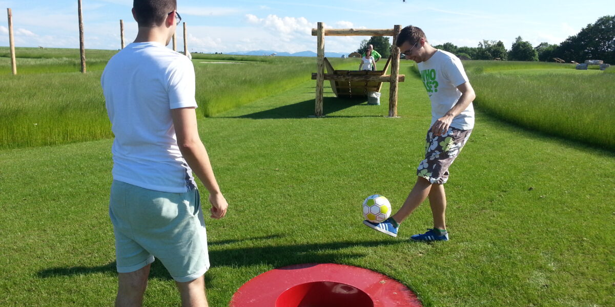 Zwei Personen spielen Fußgolf auf einer Wiese; eine von ihnen ist im Begriff, einen Fußball in ein großes rotes Loch im Boden zu schießen.