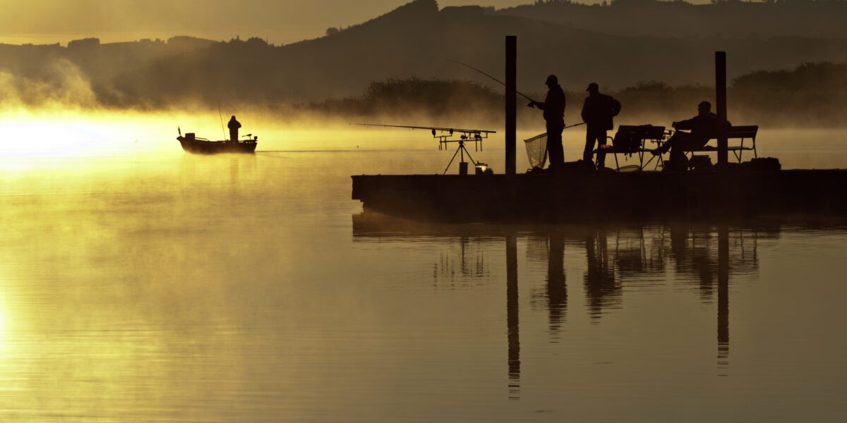 Silhouettierte Menschen angeln bei Sonnenaufgang von einem Steg aus, während Nebel von dem ruhigen See aufsteigt; ein kleines Boot ist im Hintergrund in der Nähe der entfernten Hügel zu sehen.