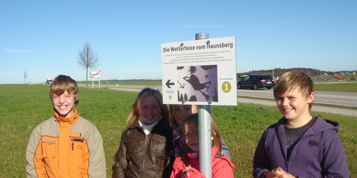 Fünf Kinder stehen an einem sonnigen Tag im Freien neben einem Schild mit der Aufschrift "Die Wetterhexe vom Haunsberg", mit blauem Himmel und grasbewachsenen Feldern im Hintergrund.