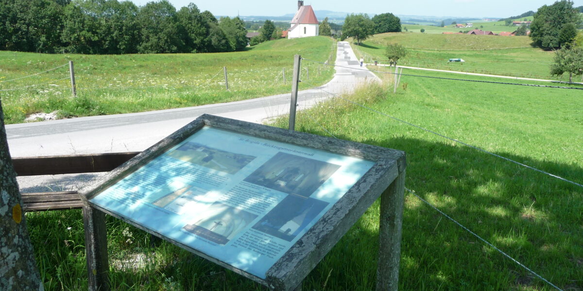 Informationsschild an einer Landstraße mit einer Kirche und Feldern im Hintergrund an einem sonnigen Tag.