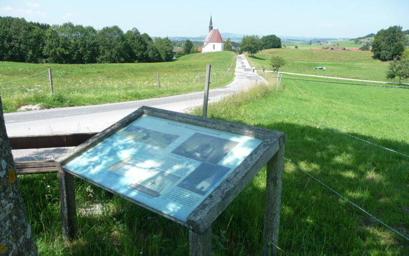Informationsschild an einer Landstraße mit einer Kirche und Feldern im Hintergrund an einem sonnigen Tag.