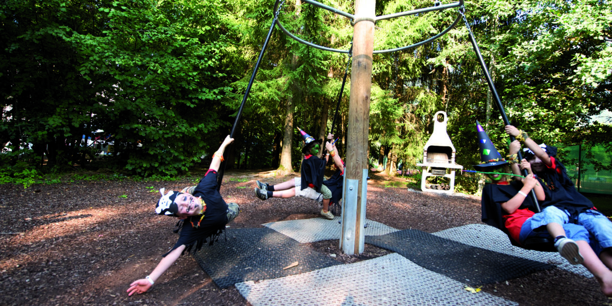 Kinder spielen auf einer drehbaren Schaukel auf einem bewaldeten Spielplatz, wobei ein Kind beim Schaukeln einen Arm nach außen streckt.