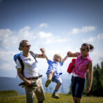 Zwei Erwachsene halten ein lächelndes Kind an den Armen und heben es spielerisch hoch, während sie an einem sonnigen Tag im Freien spazieren gehen, mit Bergen und Bäumen im Hintergrund.