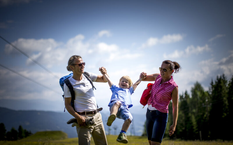 Zwei Erwachsene halten ein lächelndes Kind an den Armen und heben es spielerisch hoch, während sie an einem sonnigen Tag im Freien spazieren gehen, mit Bergen und Bäumen im Hintergrund.