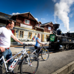 Zwei Personen mit Fahrrädern stehen auf einem Bahnsteig neben einem alten Dampfzug, mit Holzhäusern und blauem Himmel im Hintergrund.