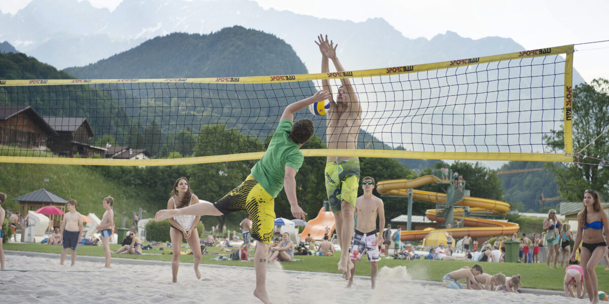 Zwei Männer springen, um einen Volleyball über ein Netz auf einem Sandplatz zu schlagen, umgeben von Zuschauern und Bergen im Hintergrund.