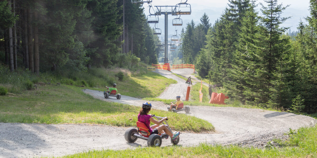 An einem sonnigen Tag fahren Menschen mit dreirädrigen Go-Karts einen gewundenen Schotterweg durch ein Waldgebiet hinunter, während über ihnen ein Skilift zu sehen ist.