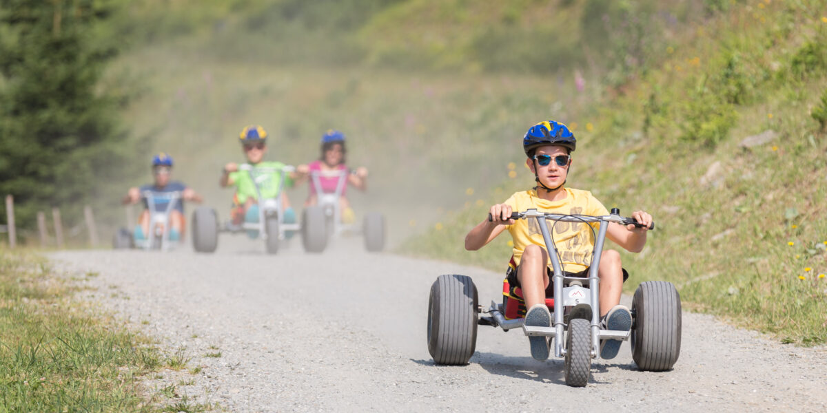 Vier Kinder mit Helmen fahren mit Gokarts auf einem Kiesweg im Freien, wobei ein Kind im Vordergrund die Gruppe anführt.