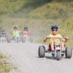 Vier Kinder mit Helmen fahren mit Gokarts auf einem Kiesweg im Freien, wobei ein Kind im Vordergrund die Gruppe anführt.