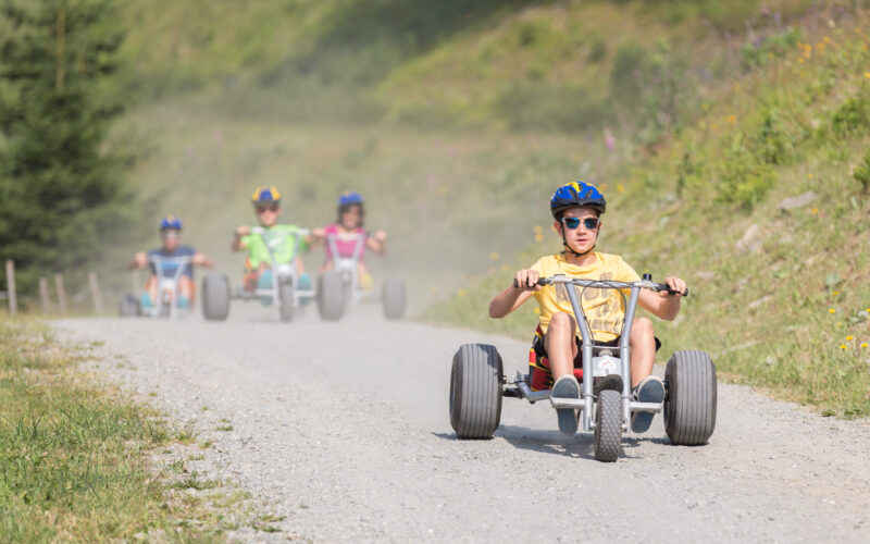 Vier Kinder mit Helmen fahren mit Gokarts auf einem Kiesweg im Freien, wobei ein Kind im Vordergrund die Gruppe anführt.