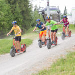 Vier Personen mit Helmen fahren mit großen Rollern einen Kiesweg in einem bewaldeten Außenbereich in der Nähe eines Skilifts hinunter.