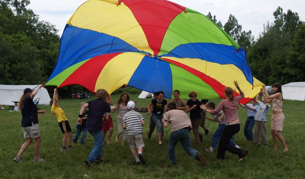 Eine Gruppe von Kindern und Erwachsenen hebt einen bunten Fallschirm in die Luft, während sie auf einer Wiese mit Bäumen und Zelten im Hintergrund spielen.