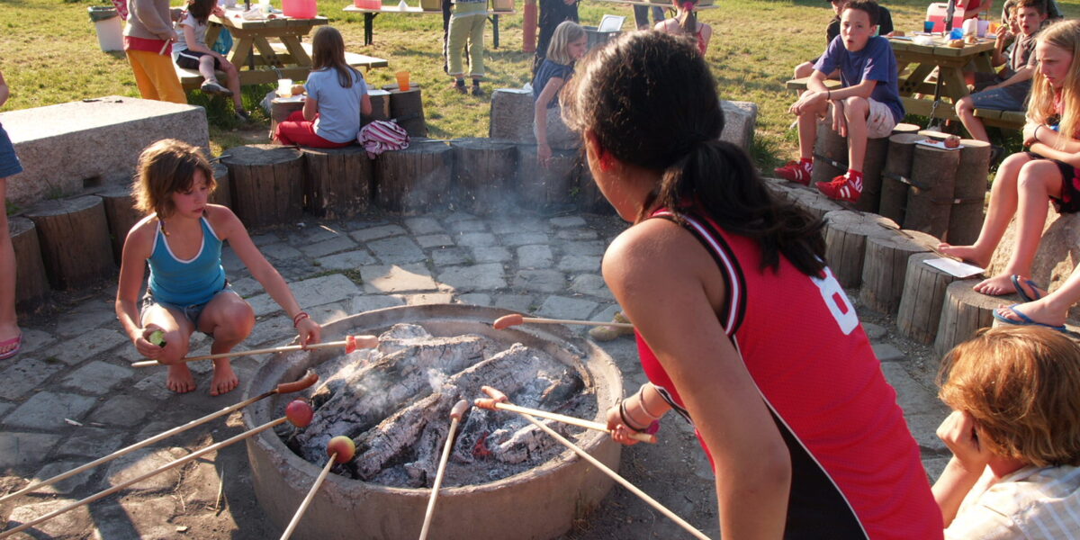 Eine Gruppe von Kindern und Erwachsenen versammelt sich an einem sonnigen Tag um eine Feuerstelle und röstet Essen auf Stöcken, während im Hintergrund Picknicktische und Aktivitäten im Freien zu sehen sind.