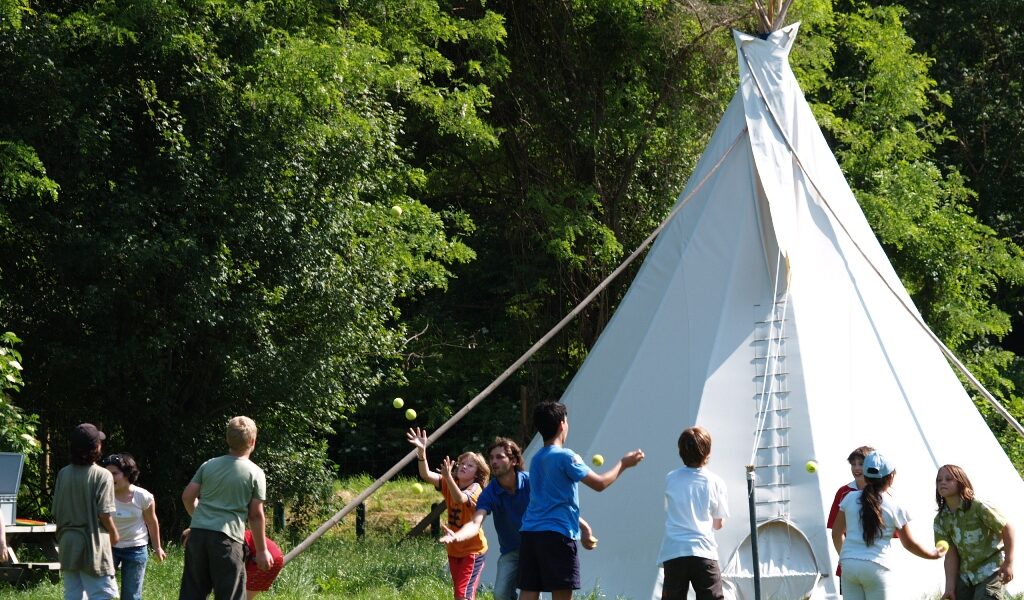 An einem sonnigen Tag spielen Kinder mit Bällen in der Nähe eines großen weißen Tipis, umgeben von grünen Bäumen und Gras.