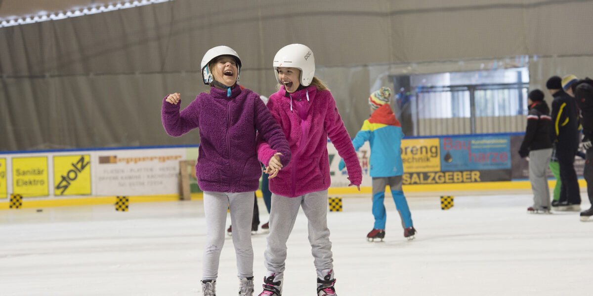 Zwei junge Mädchen, die Helme und lila Jacken tragen, laufen in einer Halle Schlittschuh, lächeln und halten sich an den Händen, während im Hintergrund andere Schlittschuhläufer zu sehen sind.