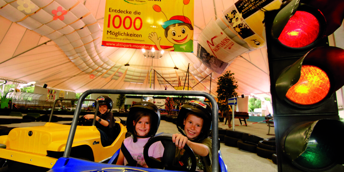 Zwei Kinder mit Helmen sitzen in einem blauen Go-Kart, während im Hintergrund ein weiteres gelbes Go-Kart, eine große Ampel und ein buntes Banner des Freizeitparks zu sehen sind.