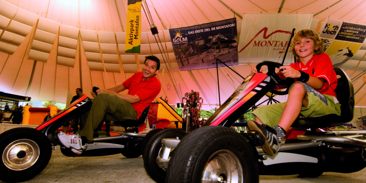 Ein Erwachsener und ein Kind fahren Gokarts unter einem großen Zelt, in dem im Hintergrund Banner hängen.