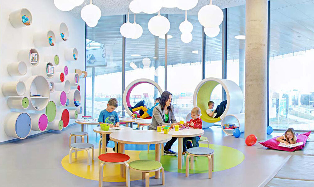 Kinder und ein Erwachsener beschäftigen sich an runden Tischen in einem hell erleuchteten, modernen Spielzimmer mit bunten Möbeln und runden Wandregalen.