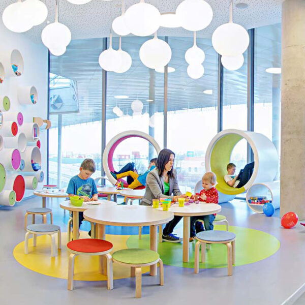 Kinder und ein Erwachsener beschäftigen sich an runden Tischen in einem hell erleuchteten, modernen Spielzimmer mit bunten Möbeln und runden Wandregalen.