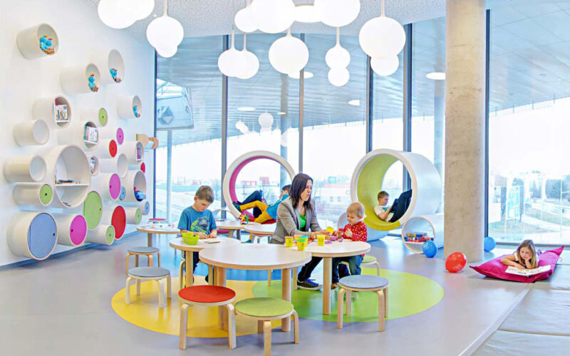 Kinder und ein Erwachsener beschäftigen sich an runden Tischen in einem hell erleuchteten, modernen Spielzimmer mit bunten Möbeln und runden Wandregalen.