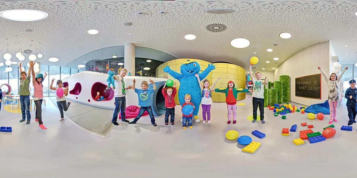 Kinder und Erwachsene stehen in einem Spielzimmer mit bunten Blöcken, einige halten Bälle, und in der Mitte posiert eine Person in einem blauen Monsterkostüm.