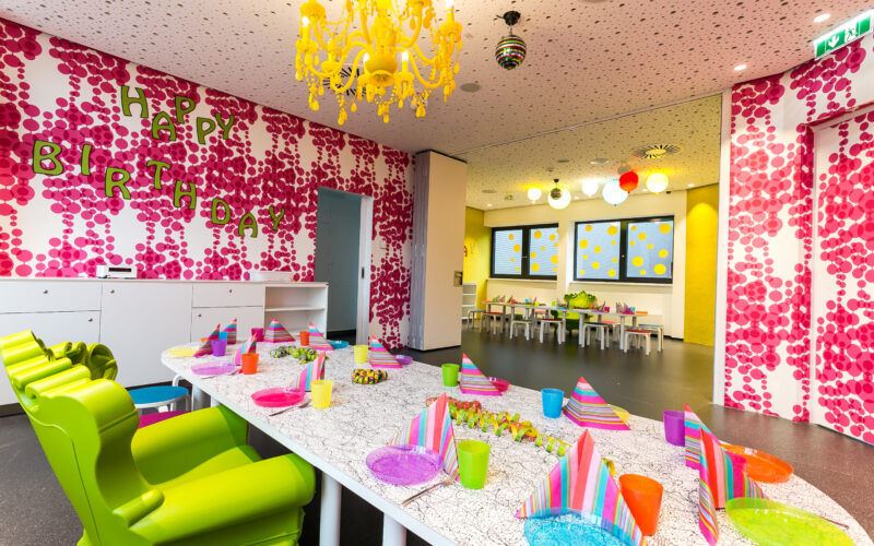 Ein farbenfroh dekorierter Raum für einen Kindergeburtstag mit bunt gedeckten Tischen, Partyhüten und einem "Happy Birthday"-Schild an der Wand.