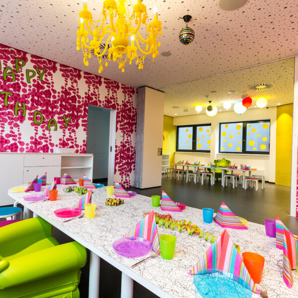 In einem bunt geschmückten Kinderpartyraum steht ein langer Tisch mit farbenfrohen Tellern, Tassen und Partyhüten, und an einer rosa-weißen Wand steht "Happy Birthday".