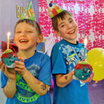 Zwei lächelnde Jungen mit Partyhüten halten Muffins mit brennenden Kerzen und einem gelben Luftballon auf einer geschmückten Geburtstagsparty.