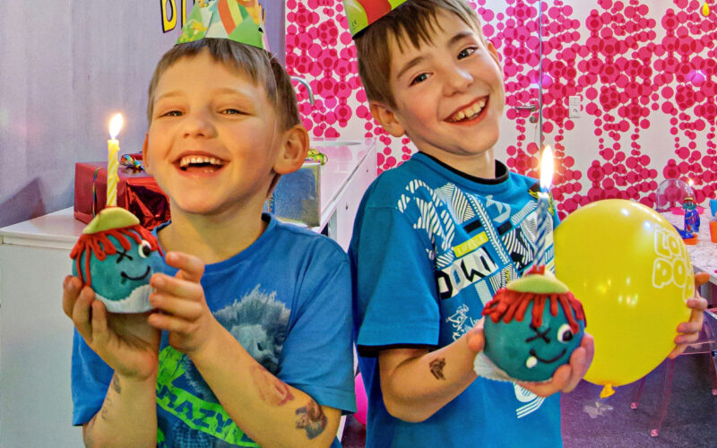 Zwei lächelnde Jungen mit Partyhüten halten Muffins mit brennenden Kerzen und einem gelben Luftballon auf einer geschmückten Geburtstagsparty.