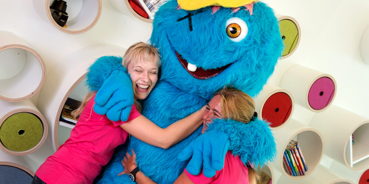 Zwei Frauen in rosafarbenen Hemden umarmen ein großes blaues Maskottchen mit gelber Mütze, das vor Regalen mit Büchern und runden Ablageflächen steht.