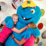 Zwei Frauen in rosa Hemden umarmen eine Person in einem großen blauen Monsterkostüm mit gelber Mütze, die vor runden Regalen mit Büchern und Spielen steht.