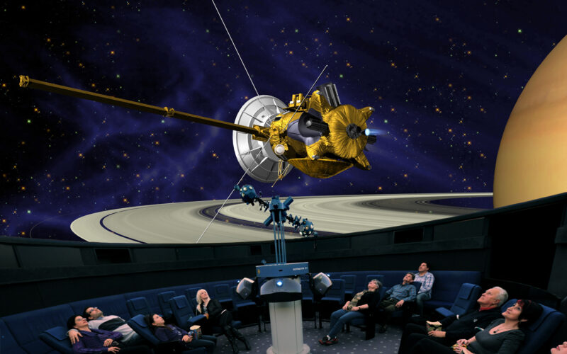 Die Menschen sitzen in einem Planetarium und blicken nach oben, während eine große Projektion eines Raumschiffs und des Saturns mit seinen Ringen an der Kuppeldecke zu sehen ist.