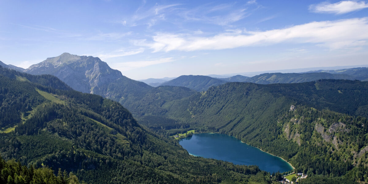 Luftaufnahme einer grün bewaldeten Berglandschaft mit einem blauen See im Tal unter einem klaren, teilweise bewölkten Himmel.