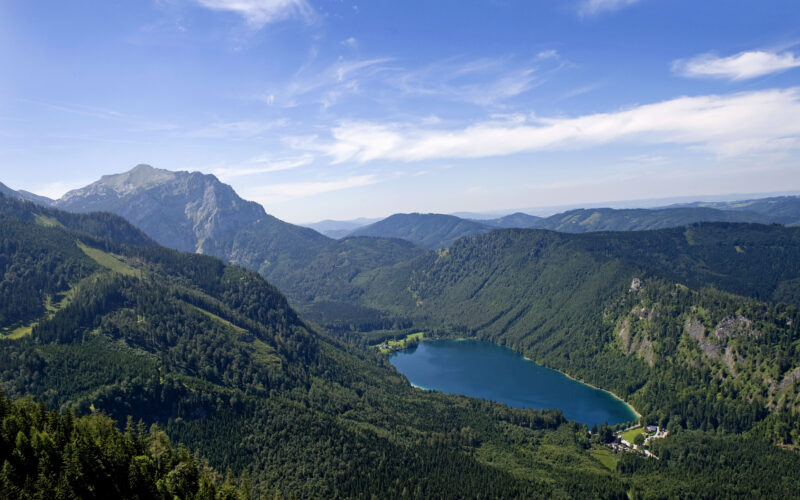Luftaufnahme einer grün bewaldeten Berglandschaft mit einem blauen See im Tal unter einem klaren, teilweise bewölkten Himmel.
