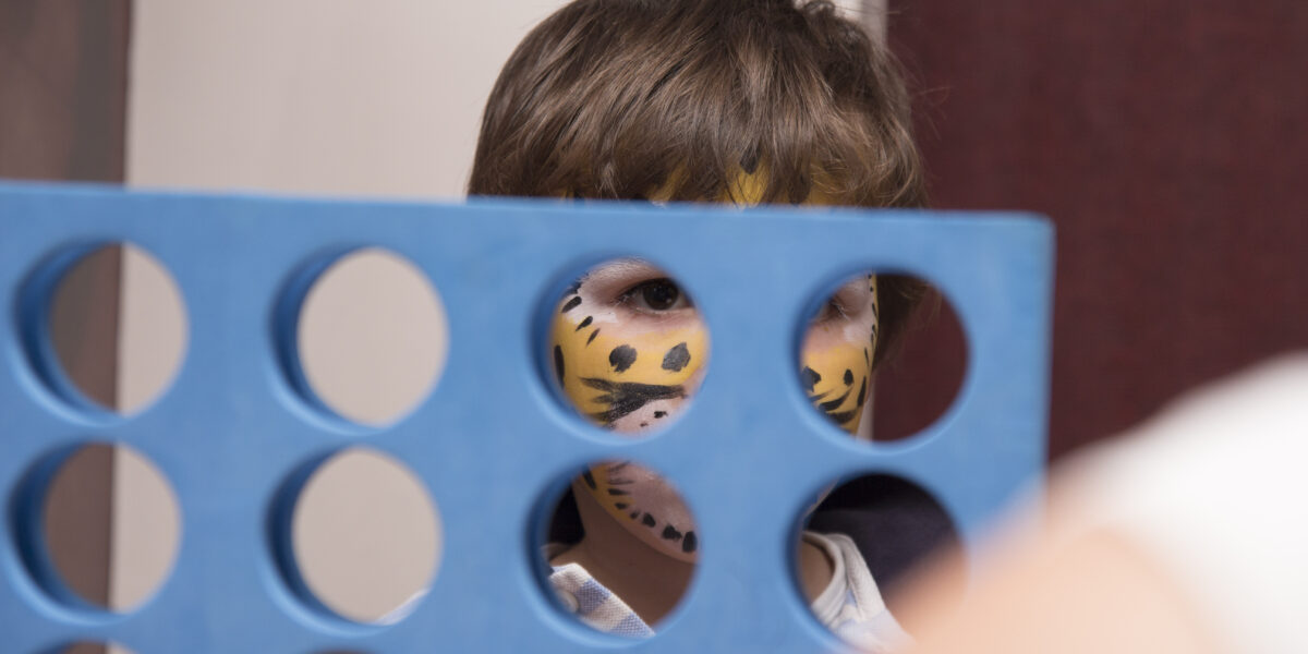Ein Kind mit bemaltem Tigergesicht schaut durch ein großes blaues Connect Four-Spielbrett.