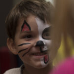 Ein kleines Kind mit braunem Haar hat ein wie ein Hund bemaltes Gesicht mit schwarzen und weißen Partien, Flecken, einer roten Zunge und schwarzen Schnurrhaaren und lächelt jemanden unscharf an.