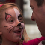 Ein junges Mädchen mit Katzengesichtsbemalung lächelt und sieht einen erwachsenen Mann an, der ihr gegenübersteht.