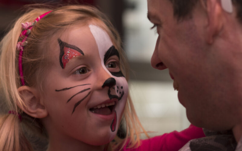 Ein junges Mädchen mit Katzengesichtsbemalung lächelt und sieht einen erwachsenen Mann an, der ihr gegenübersteht.