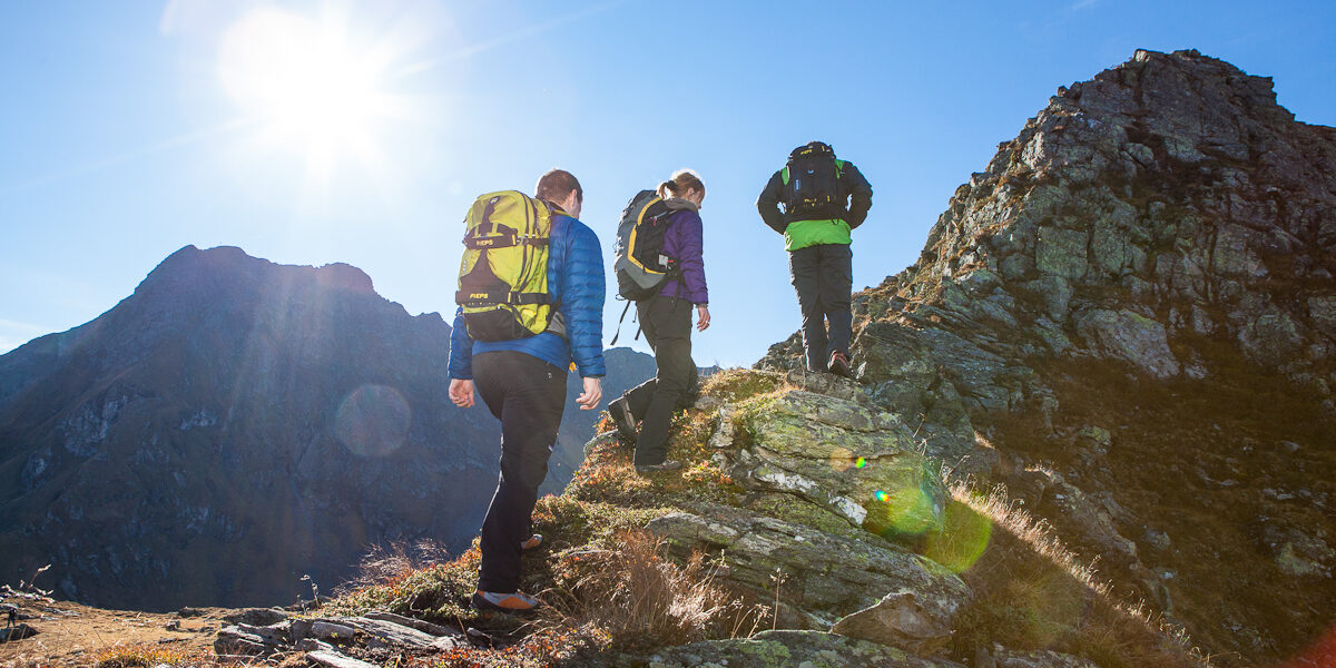 Drei Personen mit Rucksäcken wandern bei strahlendem Sonnenschein und blauem Himmel einen felsigen Bergpfad hinauf.