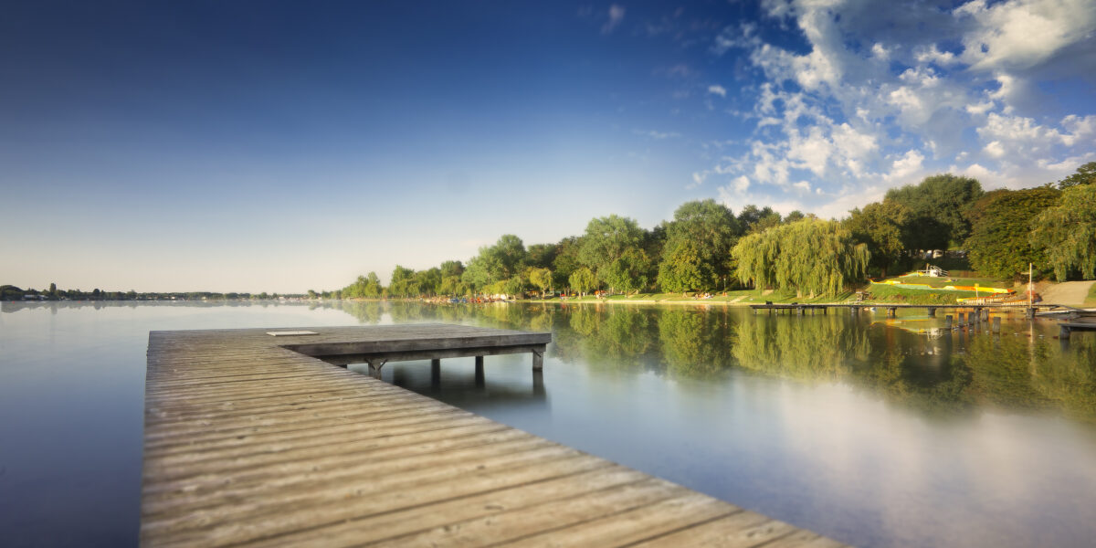 Der hölzerne Steg erstreckt sich über einen ruhigen See mit Bäumen und grasbewachsenen Ufern, die sich im Wasser spiegeln, unter einem blauen Himmel mit vereinzelten Wolken.