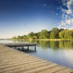 Der hölzerne Steg erstreckt sich über einen ruhigen See mit Bäumen und grasbewachsenen Ufern, die sich im Wasser spiegeln, unter einem blauen Himmel mit vereinzelten Wolken.