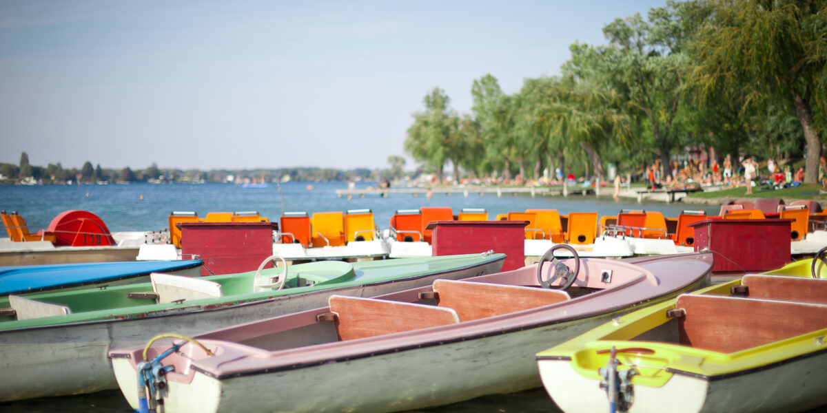 Mehrere farbenfrohe Ruder- und Tretboote liegen am Rande eines Sees vor Anker, im Hintergrund sind Bäume und Menschen am Ufer zu sehen.