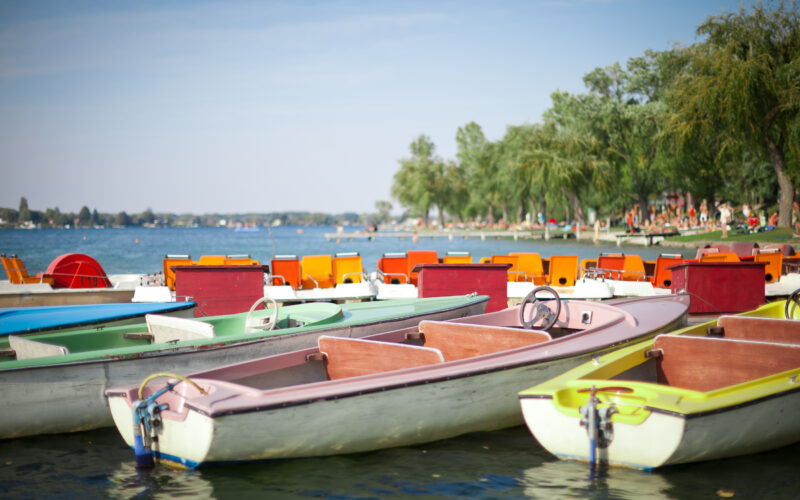 Mehrere farbenfrohe Ruder- und Tretboote liegen am Rande eines Sees vor Anker, im Hintergrund sind Bäume und Menschen am Ufer zu sehen.