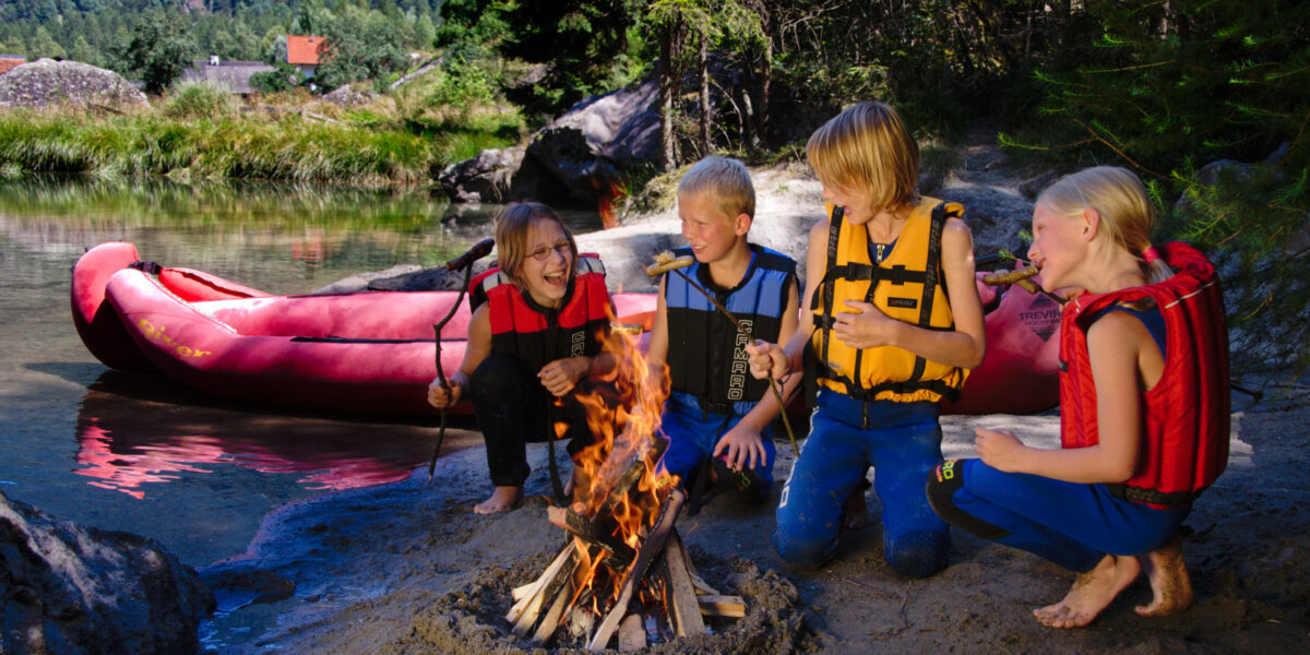 Vier Kinder mit Schwimmwesten rösten Marshmallows über einem Lagerfeuer an einem sandigen Flussufer mit einem roten Schlauchboot und Bäumen im Hintergrund.