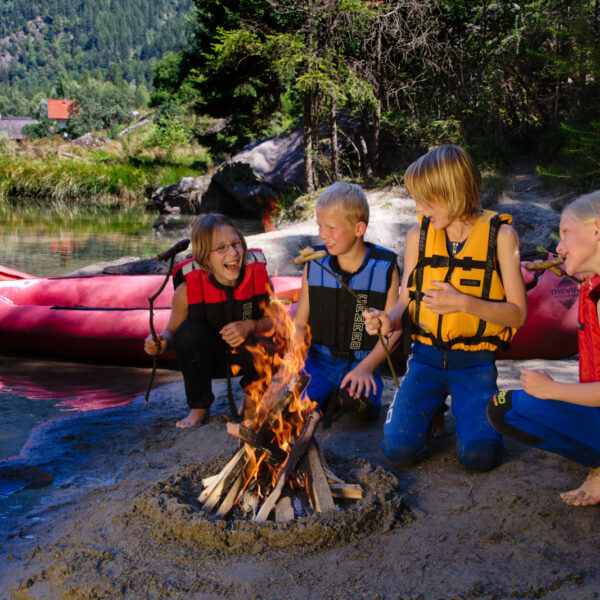 Vier Kinder mit Schwimmwesten rösten Marshmallows über einem Lagerfeuer an einem sandigen Flussufer mit einem roten Schlauchboot und Bäumen im Hintergrund.