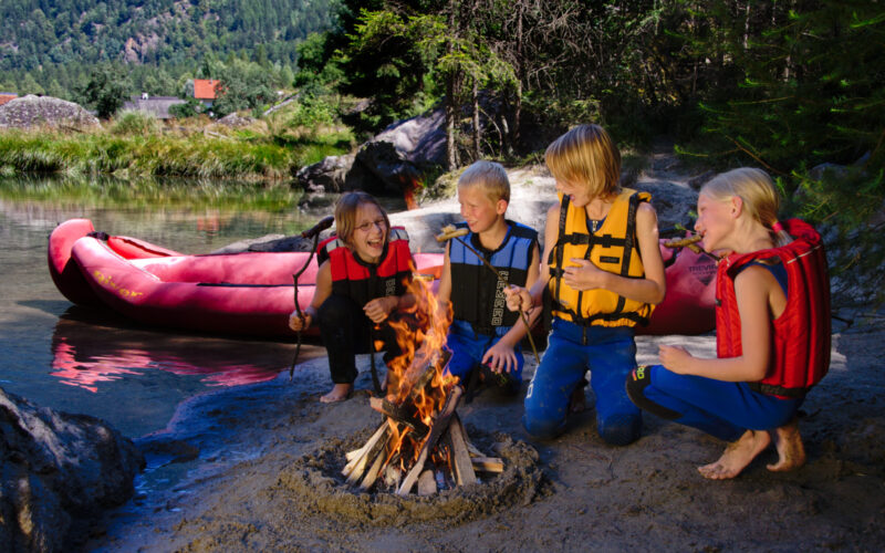 Vier Kinder mit Schwimmwesten rösten Marshmallows über einem Lagerfeuer an einem sandigen Flussufer mit einem roten Schlauchboot und Bäumen im Hintergrund.